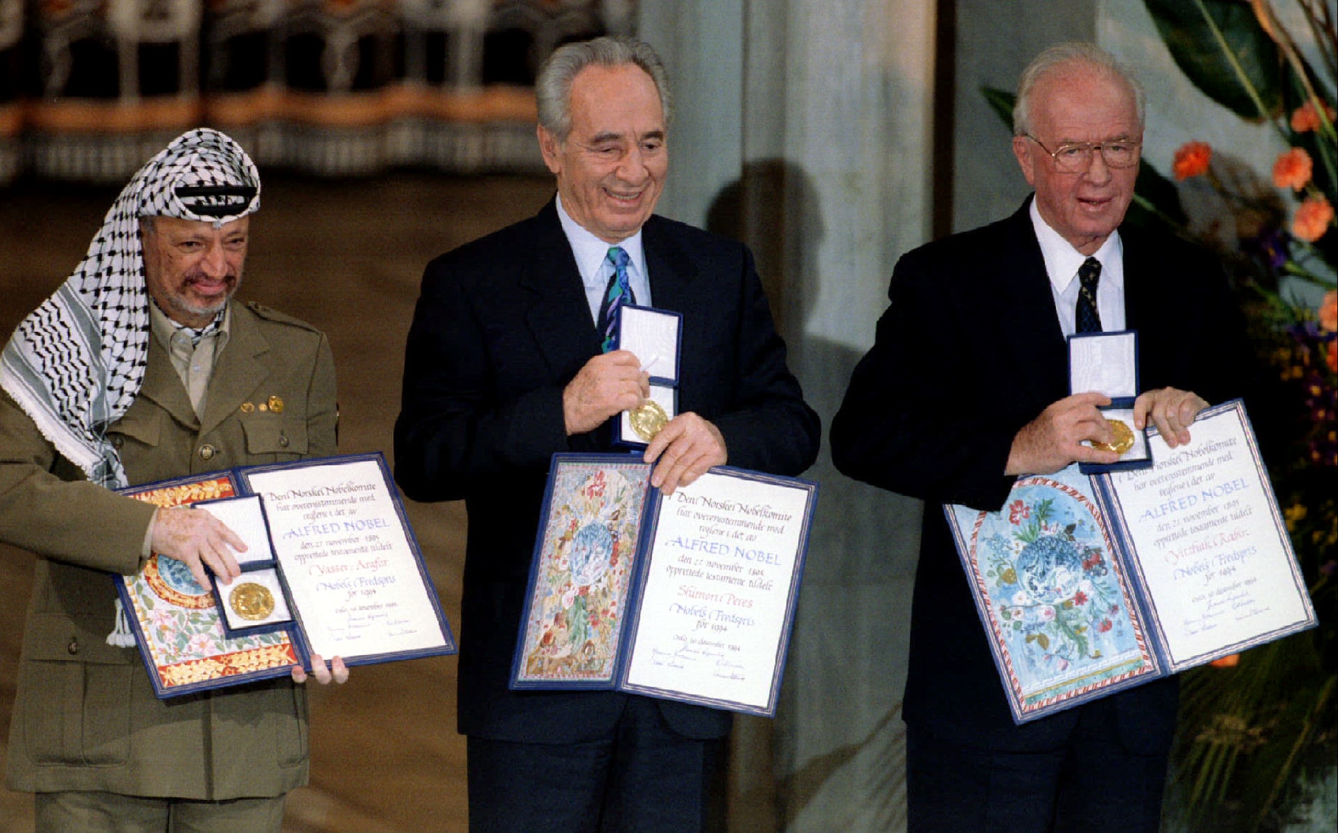 Three men stand together posing for a photo holding a medal and a certificate each.