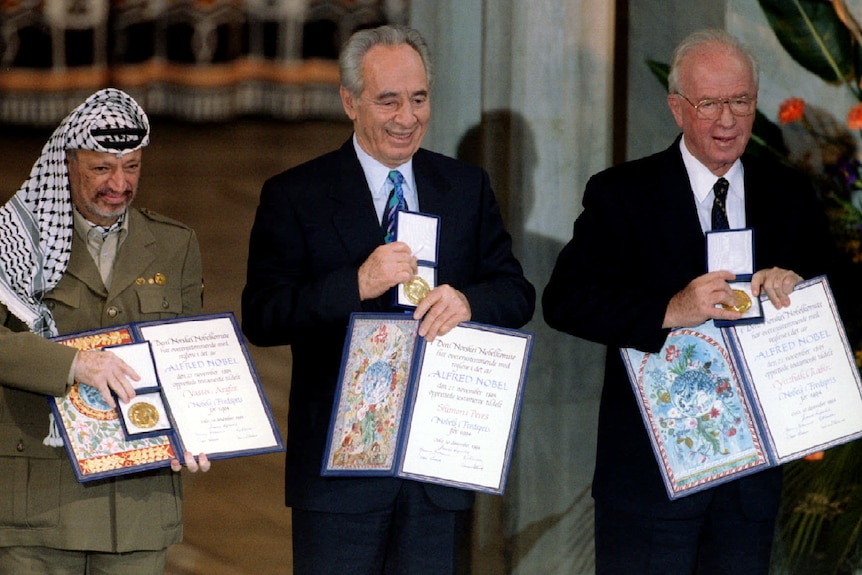 Three men stand together posing for a photo holding a medal and a certificate each.