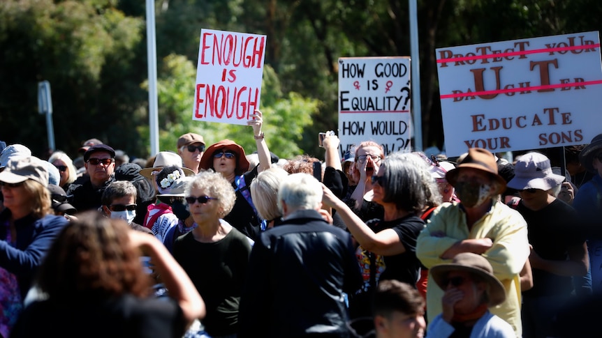 A crowd of mostly women gather outside, holding signs, one reads ENOUGH IS ENOUGH in red text