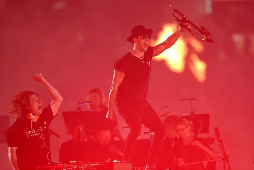 Performer Timmy Trumpet stands on a step waving a trumpet in the air.  He seems to be dancing and the lighting is red