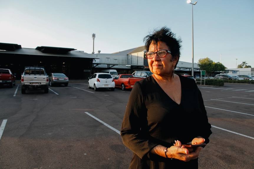 A woman wearing glasses stands holding her mobile, looking to one side. Behind her is a shopping centre and its carpark.