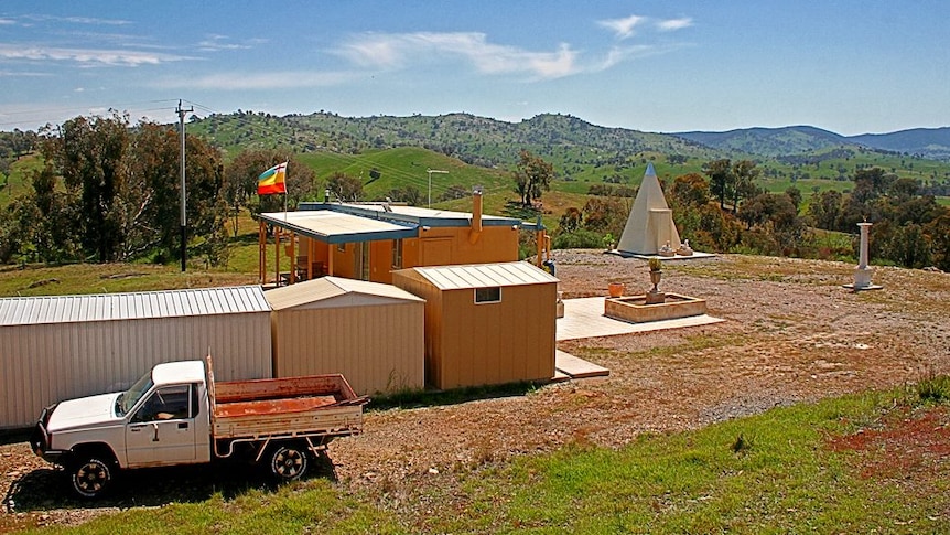 A high shot showing buildings and a 4WD on a rural property near mountains and under a blue sky.