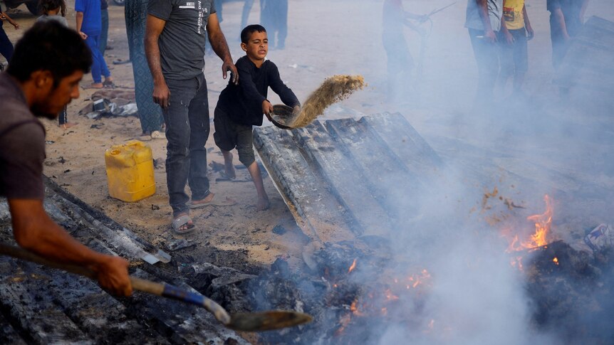 A small boy thrown sand onto a fire using a pan, with a man doing the same using a shovel in the foreground
