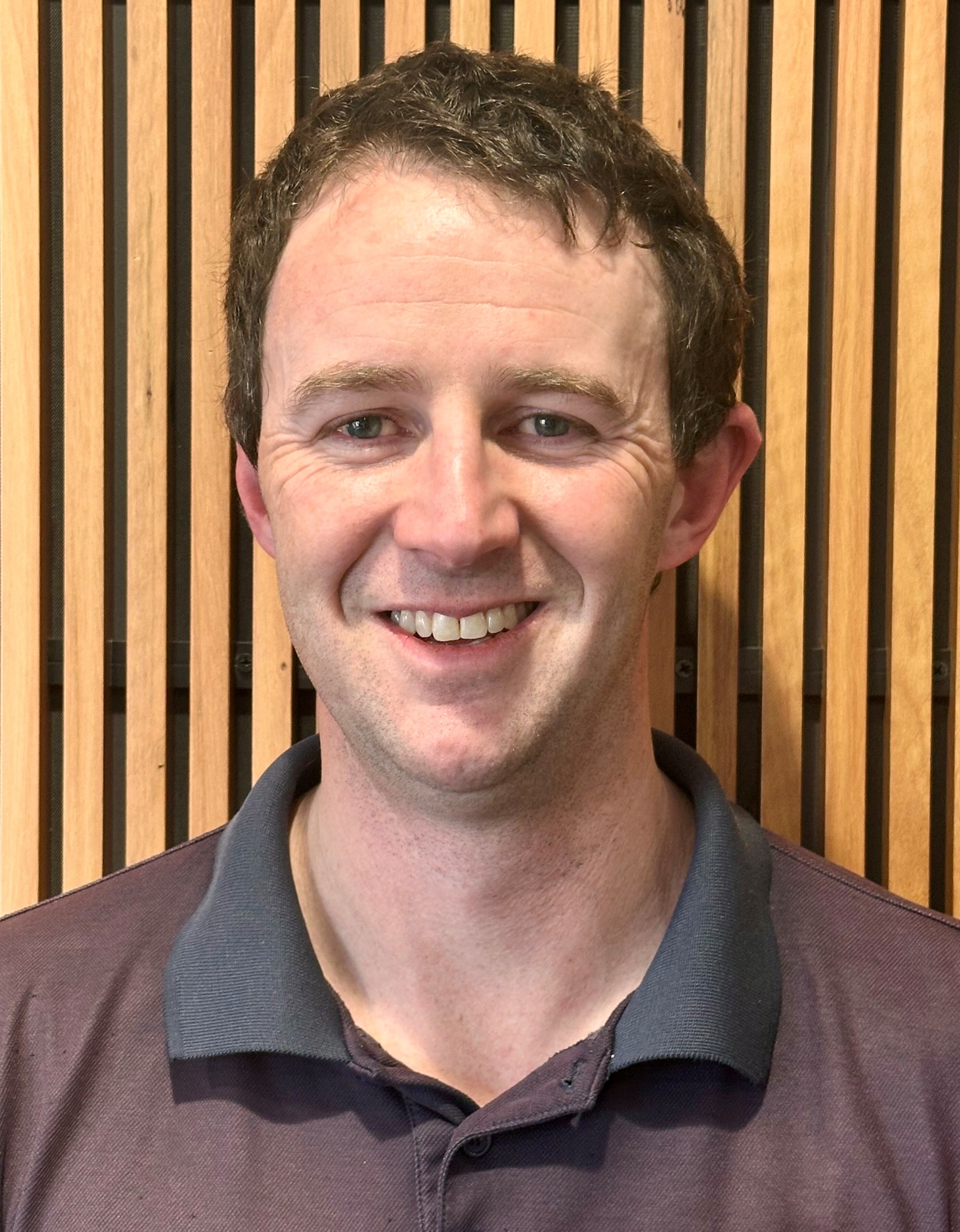 A smiling man with short hair stands in front of a wood-panelled wall.