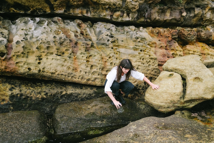 A brown haired 30-something woman squats down to examine sandstone rocks.
