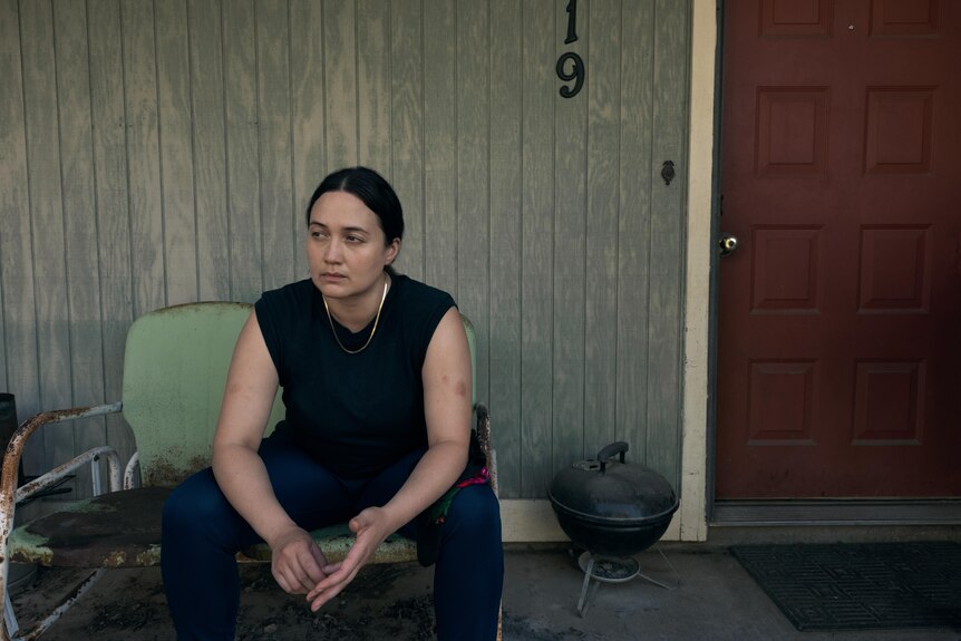 A butch woman in a black tank top sits on a stoop staring into the distance. 