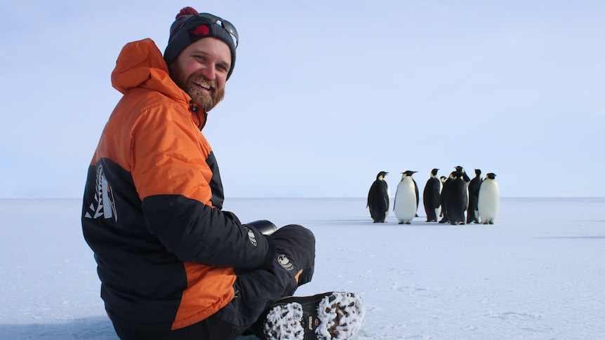 a man in snow gear is sitting cross legged on the snow, a group of penguins are in front of him