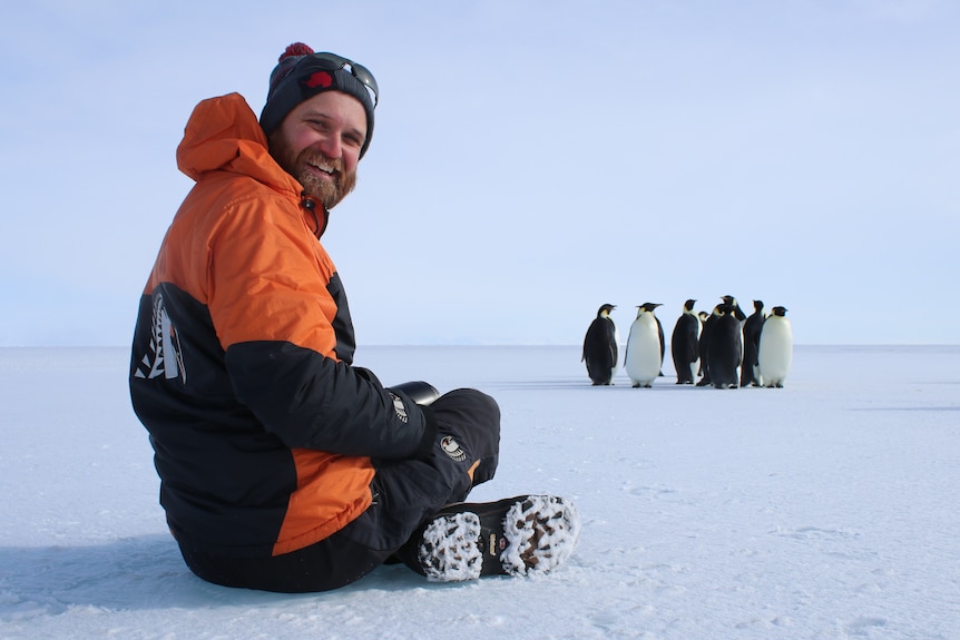 a man in snow gear is sitting cross legged on the snow, a group of penguins are in front of him
