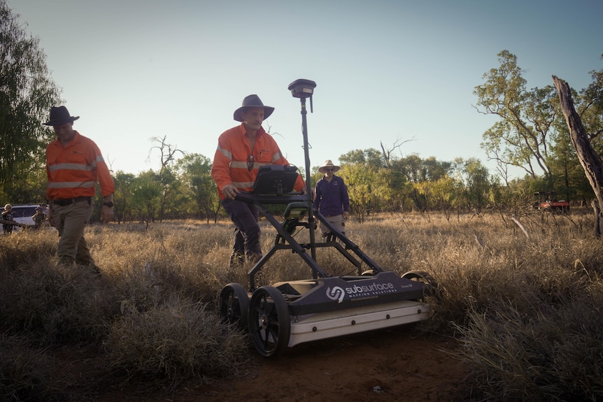 A man pushes a lawnmower like device across rough ground with others watching on