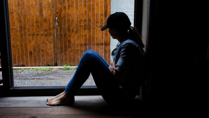 A young woman is silhouetted by a window, wearing a cap and hugging herself.