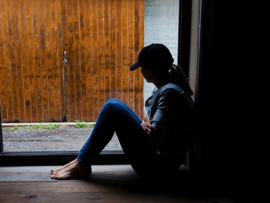 A young woman is silhouetted by a window, wearing a cap and hugging herself.