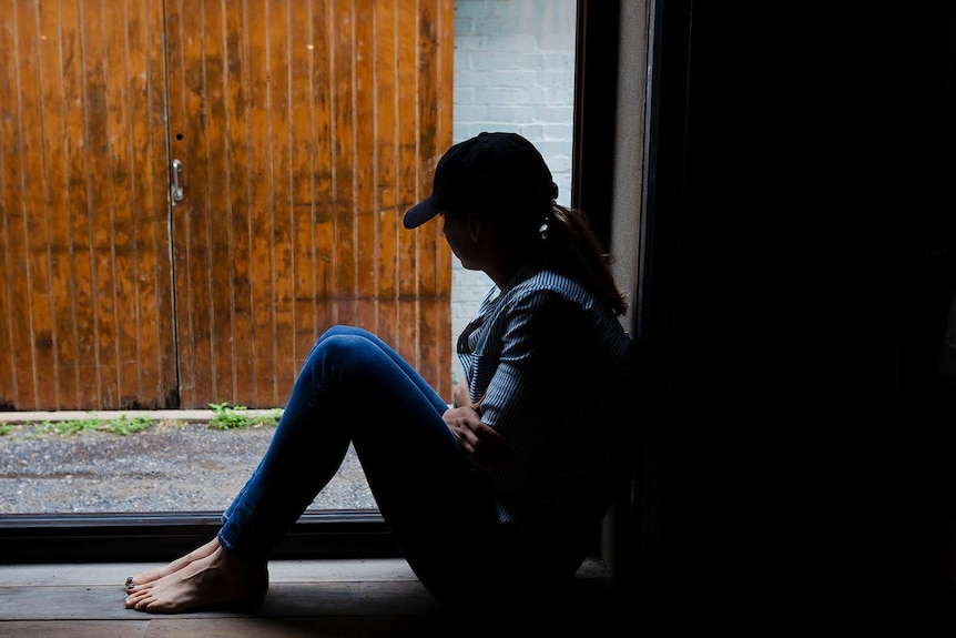 A young woman is silhouetted by a window, wearing a cap and hugging herself.