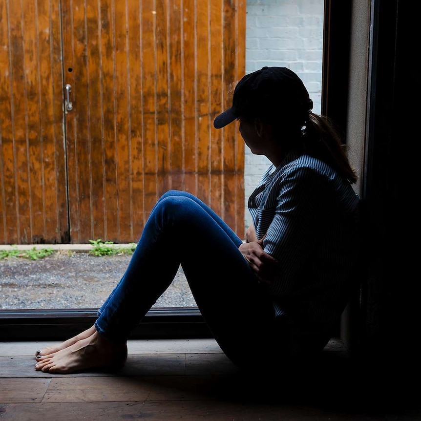 A young woman is silhouetted by a window, wearing a cap and hugging herself.