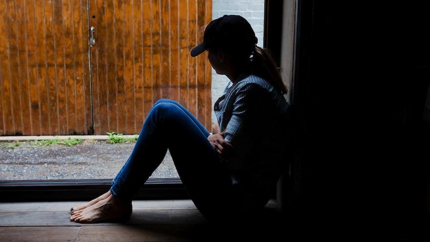 A young woman is silhouetted by a window, wearing a cap and hugging herself.