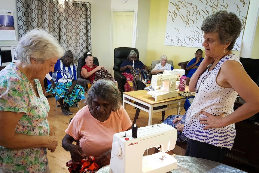 An Indigenous woman sits at a sewing machine, with a woman standing either side of her and other women sitting in the background