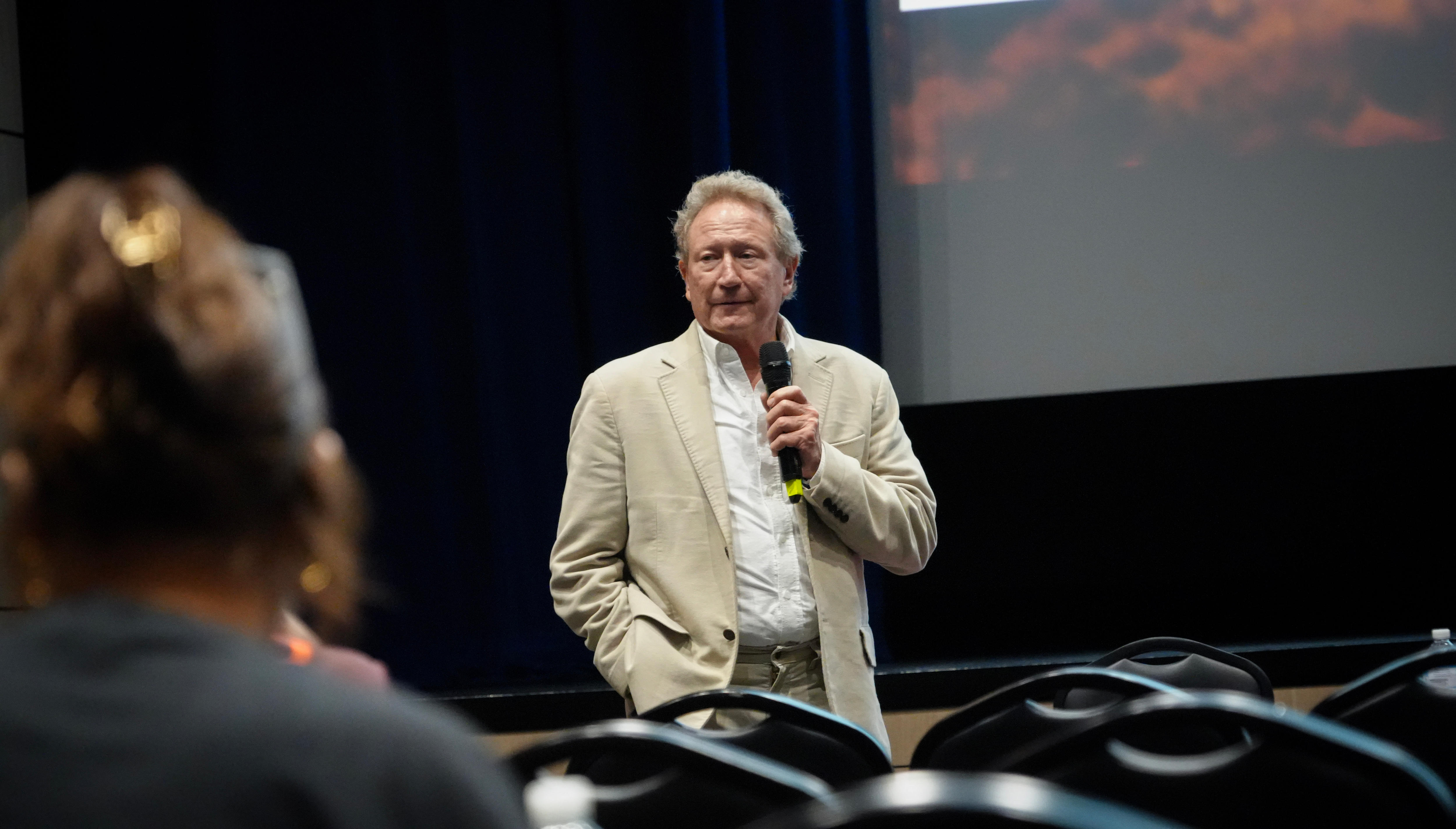 Andrew Forrest speaks at the front of an auditorium with a microphone in his hand and wears a beige suit.