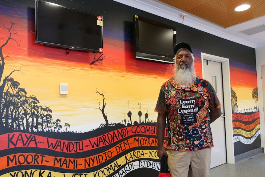 An Aboriginal man stands in front of red, black and yellow mural.