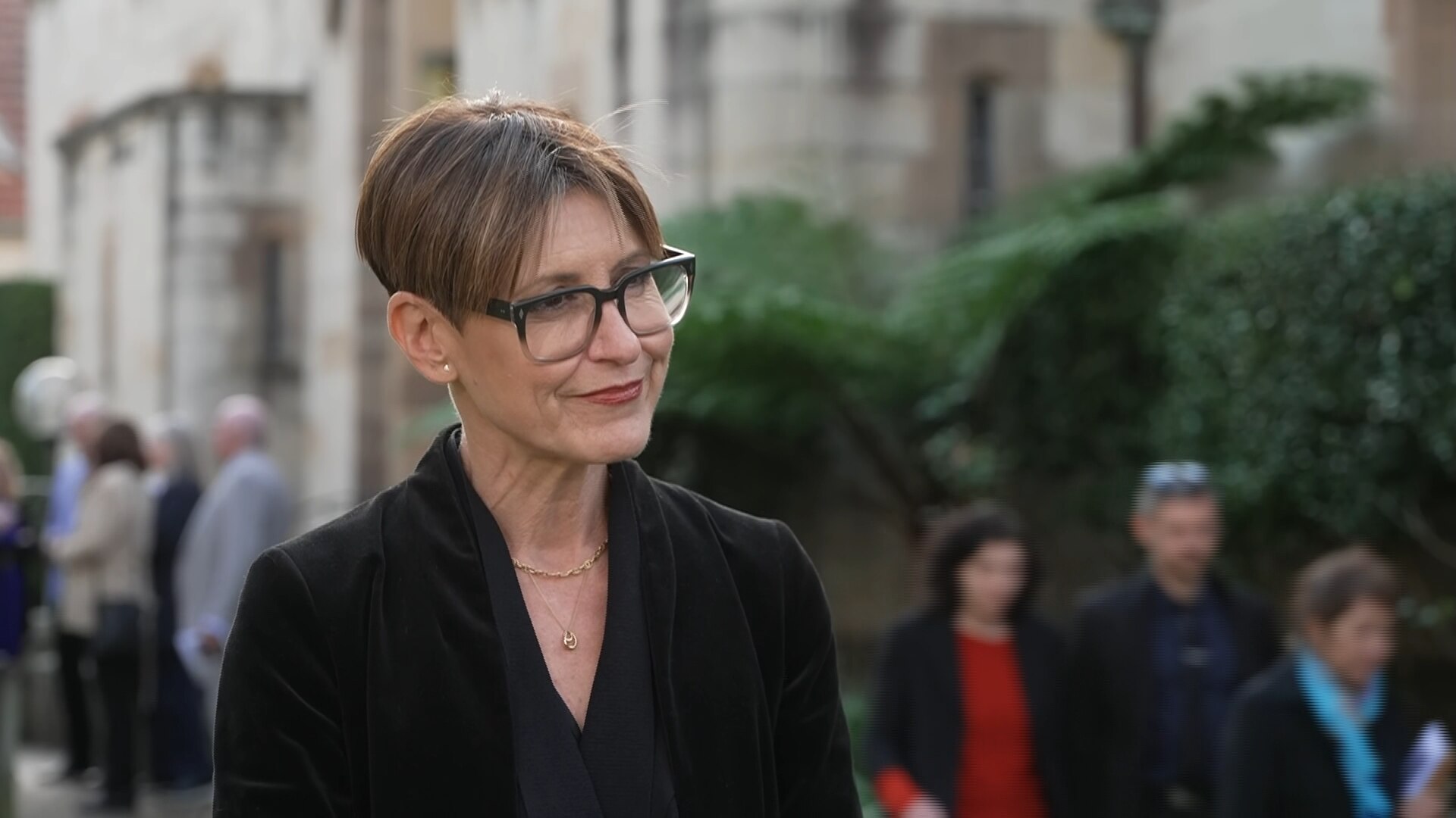 A middleaged woman with a brown bob, wearing a black blazer, stands outside a church.