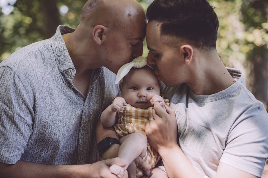 Two dads holding their baby daughter between them and, with eyes closed, lovingly kissing her on the head.