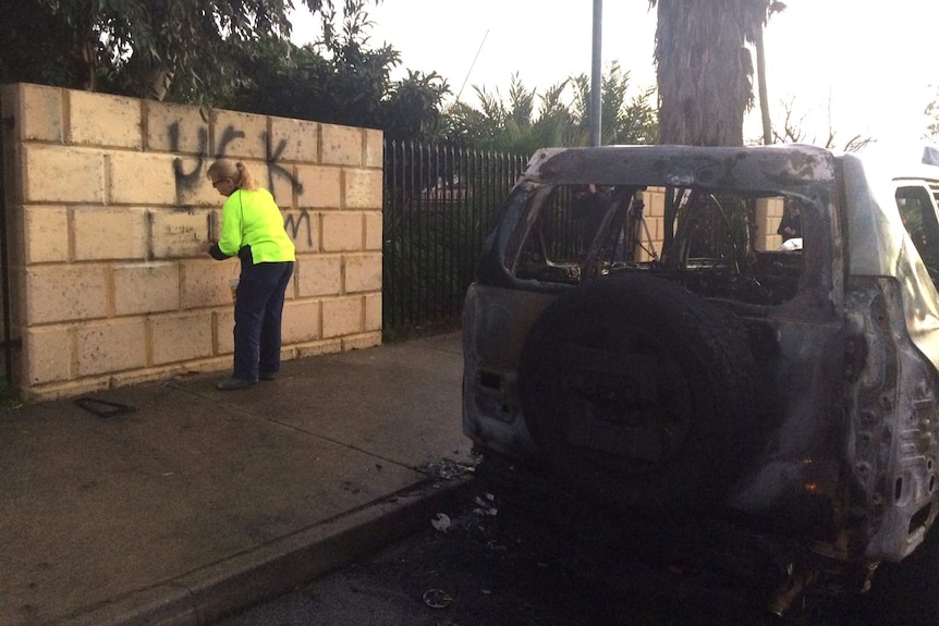 A woman cleans anti-Islam graffiti off a wall in front of a burnt-out four-wheel-drive.