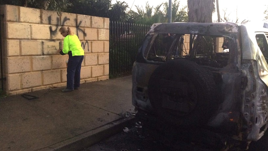 A woman cleans anti-Islam graffiti off a wall in front of a burnt-out four-wheel-drive.