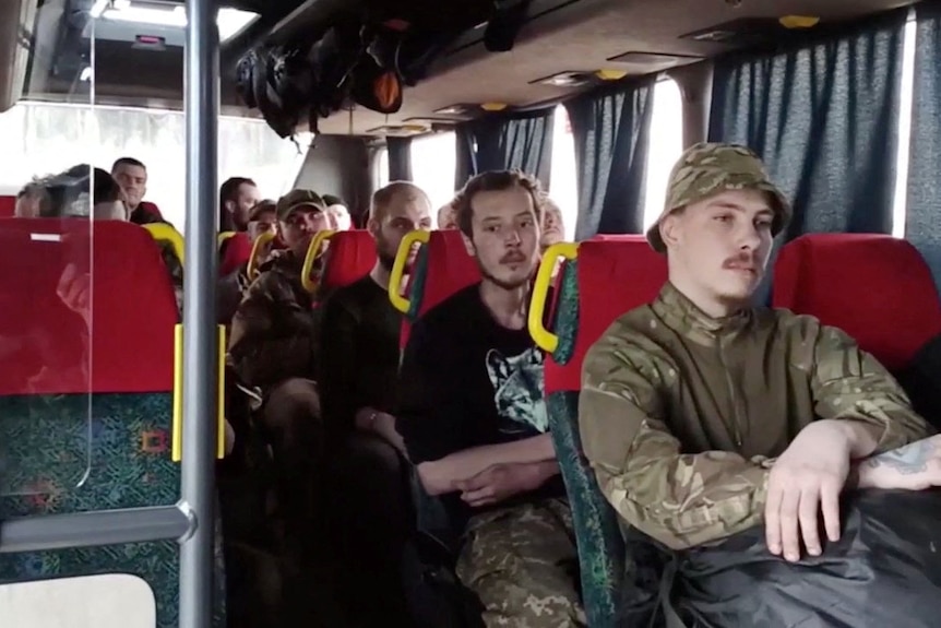 Men sit on a bus in army uniform with expressionless faces.
