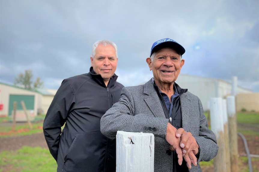 Two men stand in front of poles under a cloudy sky and green grass. Man in front elderly, wears cap, smiles.