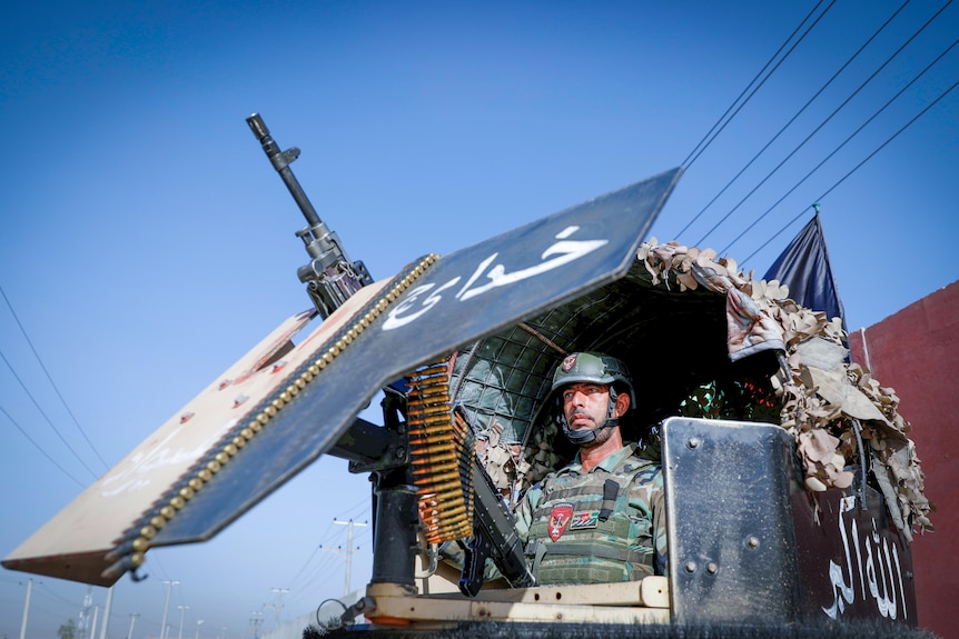 A man in combat gear looks out the window of a military vehicle with a huge machine gun attached 