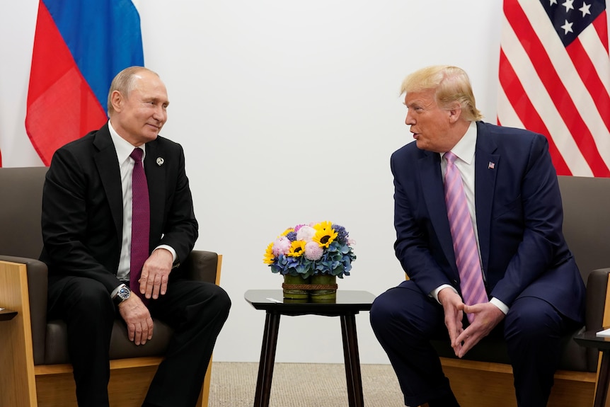An older Russian man smiles at an older American man as they sit in armchairs in front of their flags.