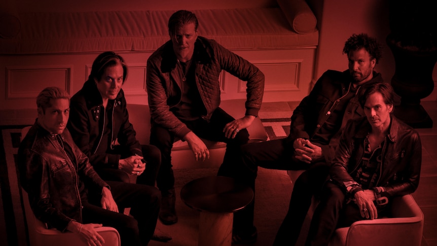 A red filter over a portrait of the five members of Queens Of The Stone Age lounging in a room