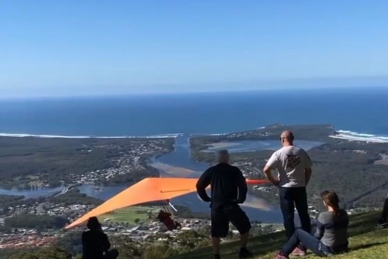 A hang glider takes off from the top of a coastal mountain, bushland, a river and the ocean in the distance.