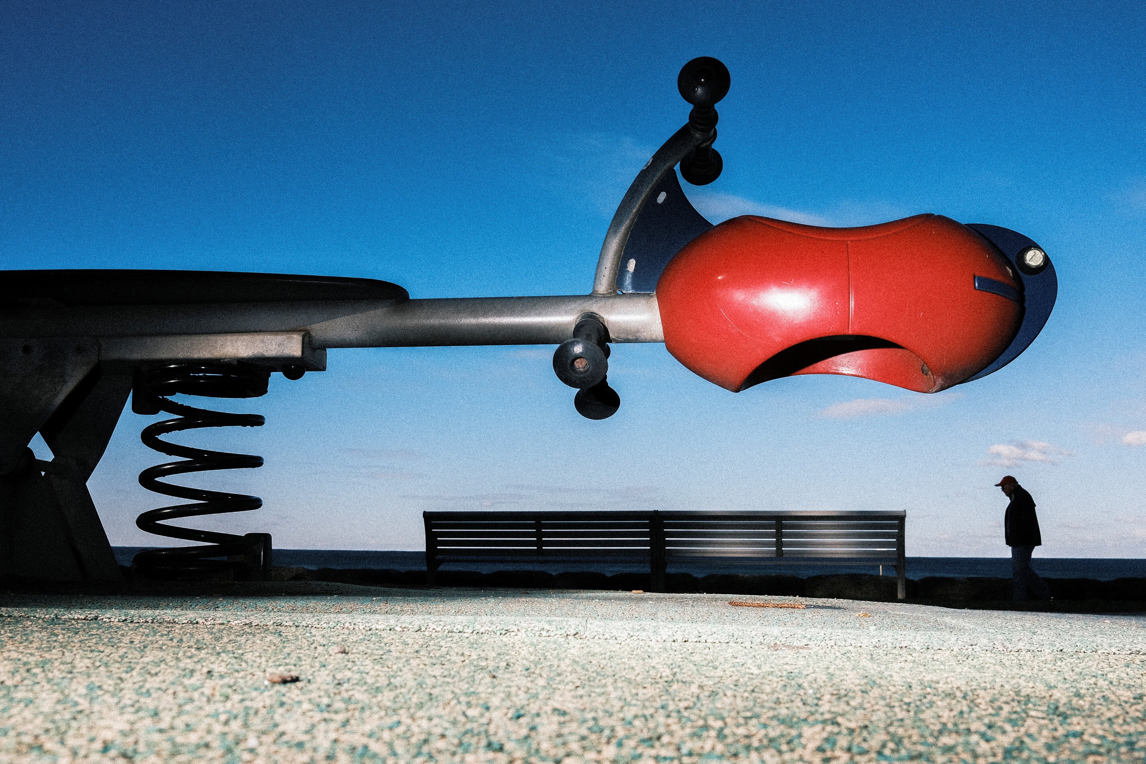 A low wide shot of a seesaw dwarfing a silhouetted person in the background walking along a path near a beach.