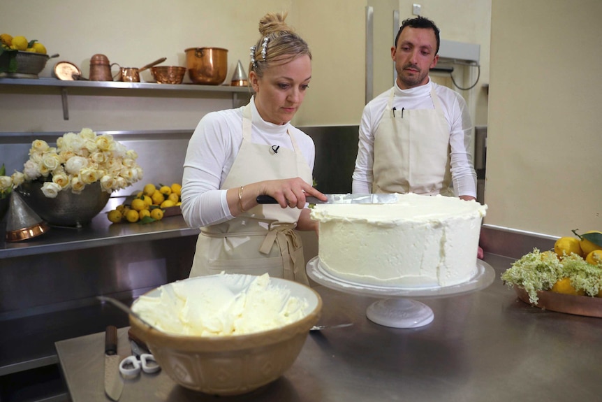 A baker finishes icing a tier of a cake, while another baker watches on.