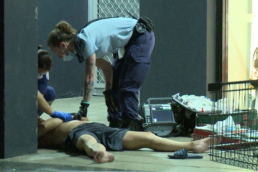 A police officer and an ambulance officer tend to a man lying on the ground with his legs splayed. 