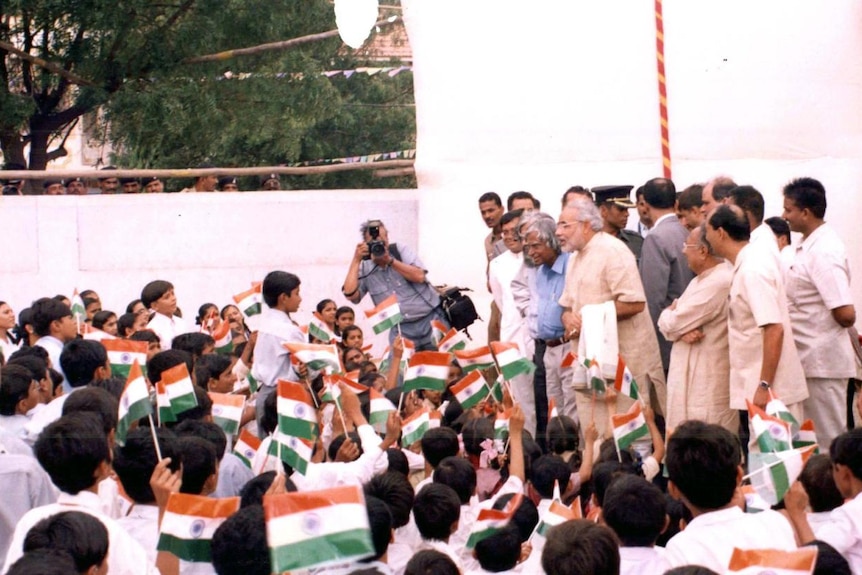 Narendra Modi stands on stage surrounded by people, speaking to a crowd waving small Indian flags