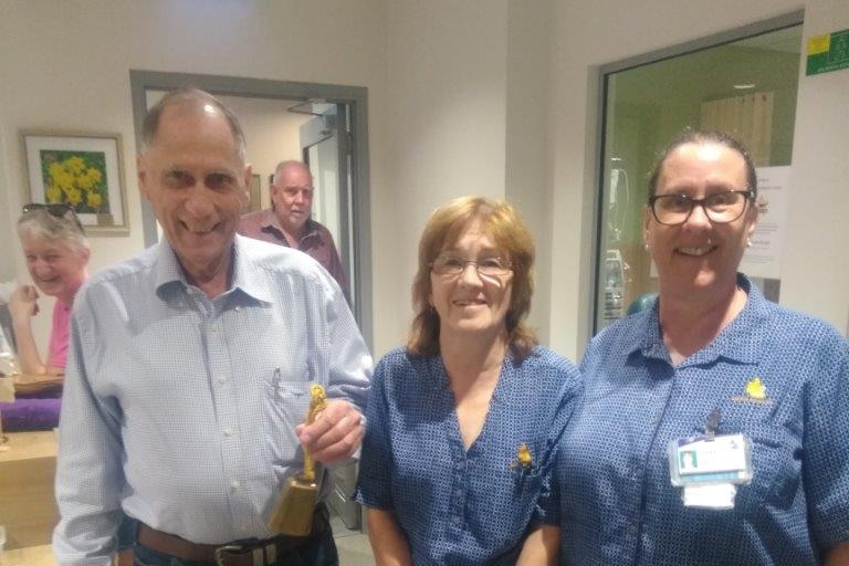 An older man smiles, he is holding a bell. There are two nurses beside him. 