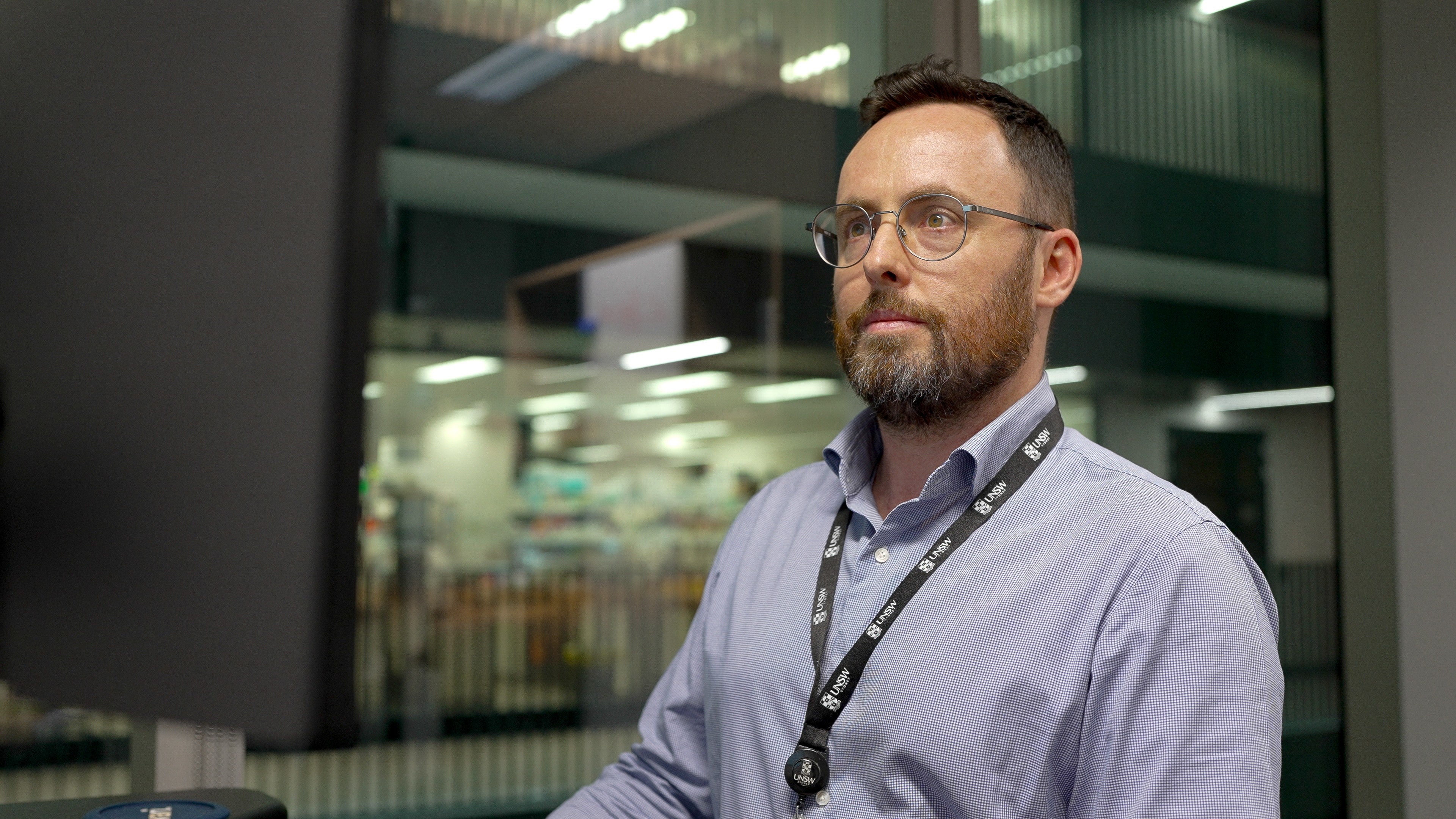 Martin Smith in a blue long sleeve and lanyard, glasses, sitting at a desk and looking at a laptop monitor.