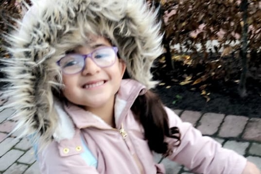 A young girl in bright purple-rimmed glasses and a fur-lined jacket is cycling on a pink bike.