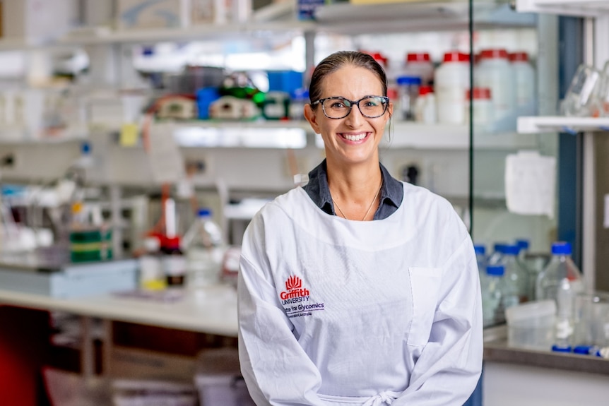 A woman with spectacles in the lab wearing a white coat and smiling.