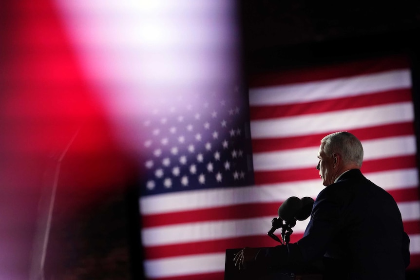 A man with white hair speaks at a podium with a US flag in front of him.