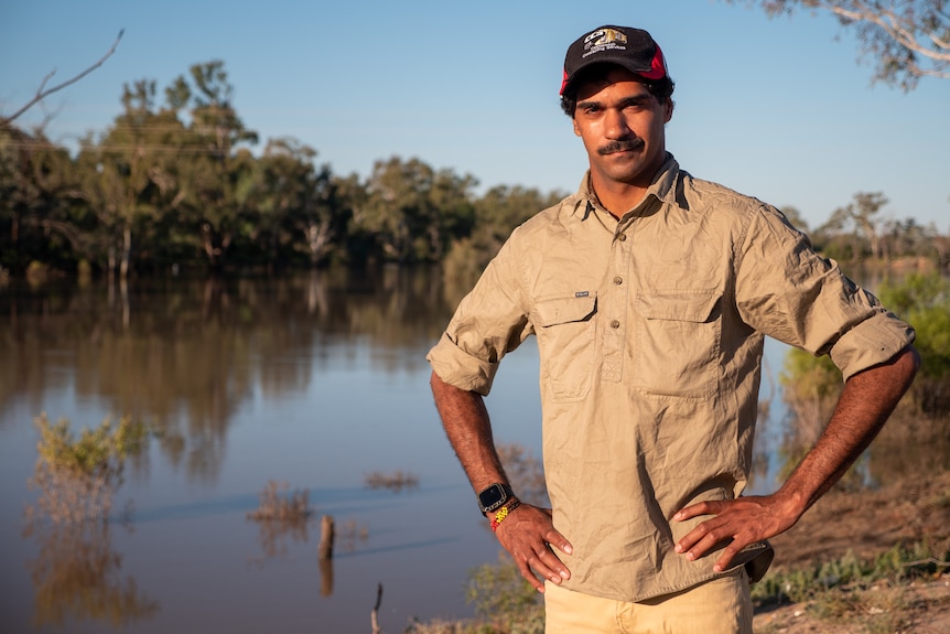 A man wearing khaki clothes and a cap stands near a waterway. 