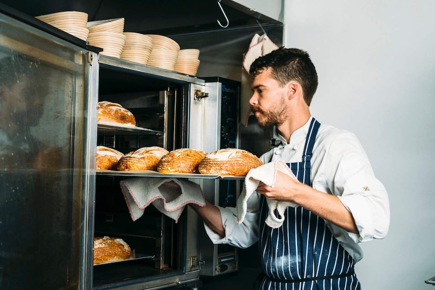 A baker takes a tray of bread loaves out of an oven