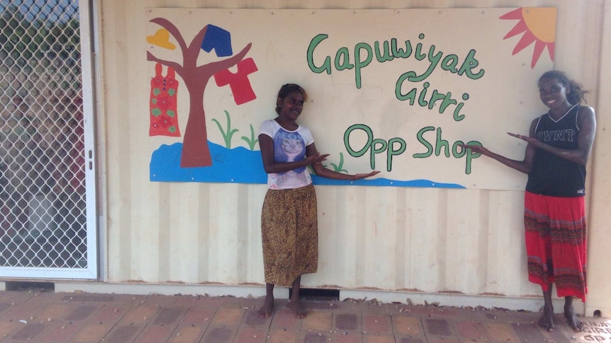 Two teenage girls stand in front of a sign reading "Gupuwiyak Girri opp shop".