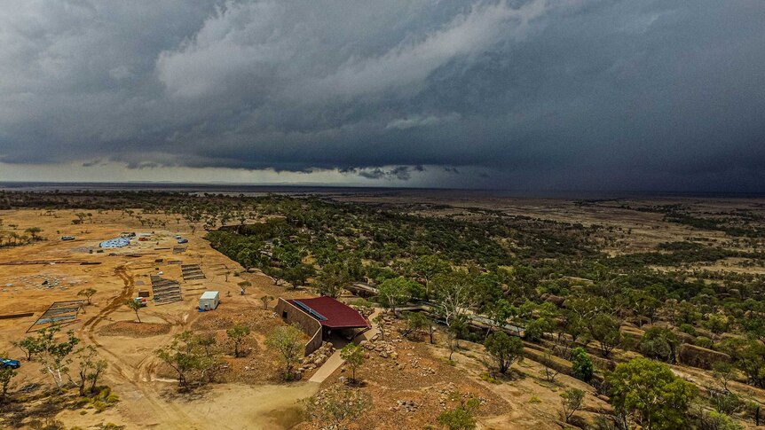 A drone shot shows dark clouds rolling in across the sky over the outback landscape.