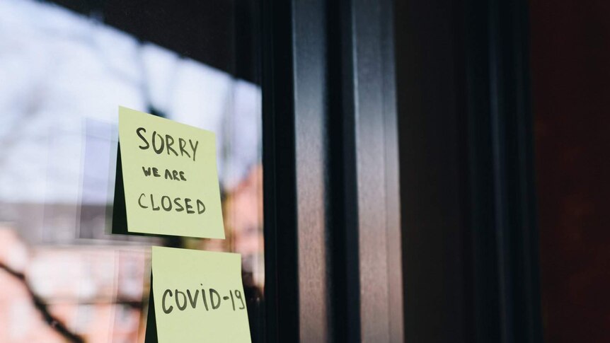 Two sticky notes, with the words "Sorry we are closed, COVID-19", are stuck to a shop window, which also reflects trees.