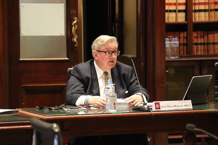 An older, bespectacled man in a dark suit sits at a desk.