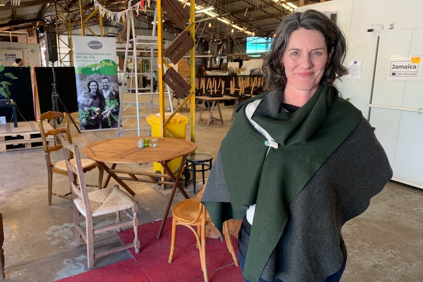 A woman with dark hair and blue eyes stands in a warehouse.