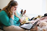 A woman looks puzzled at a laptop, sitting on a bed with her dog