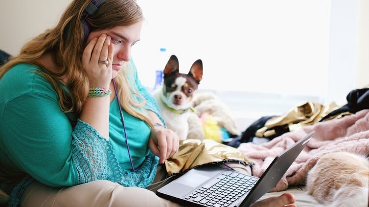 A woman looks puzzled at a laptop, sitting on a bed with her dog