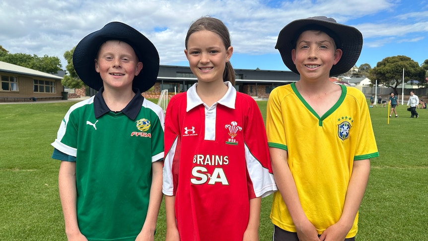 Young kids, a boy on the left in a green jersey, a girl in a red jersey and a boy on the right in yellow.